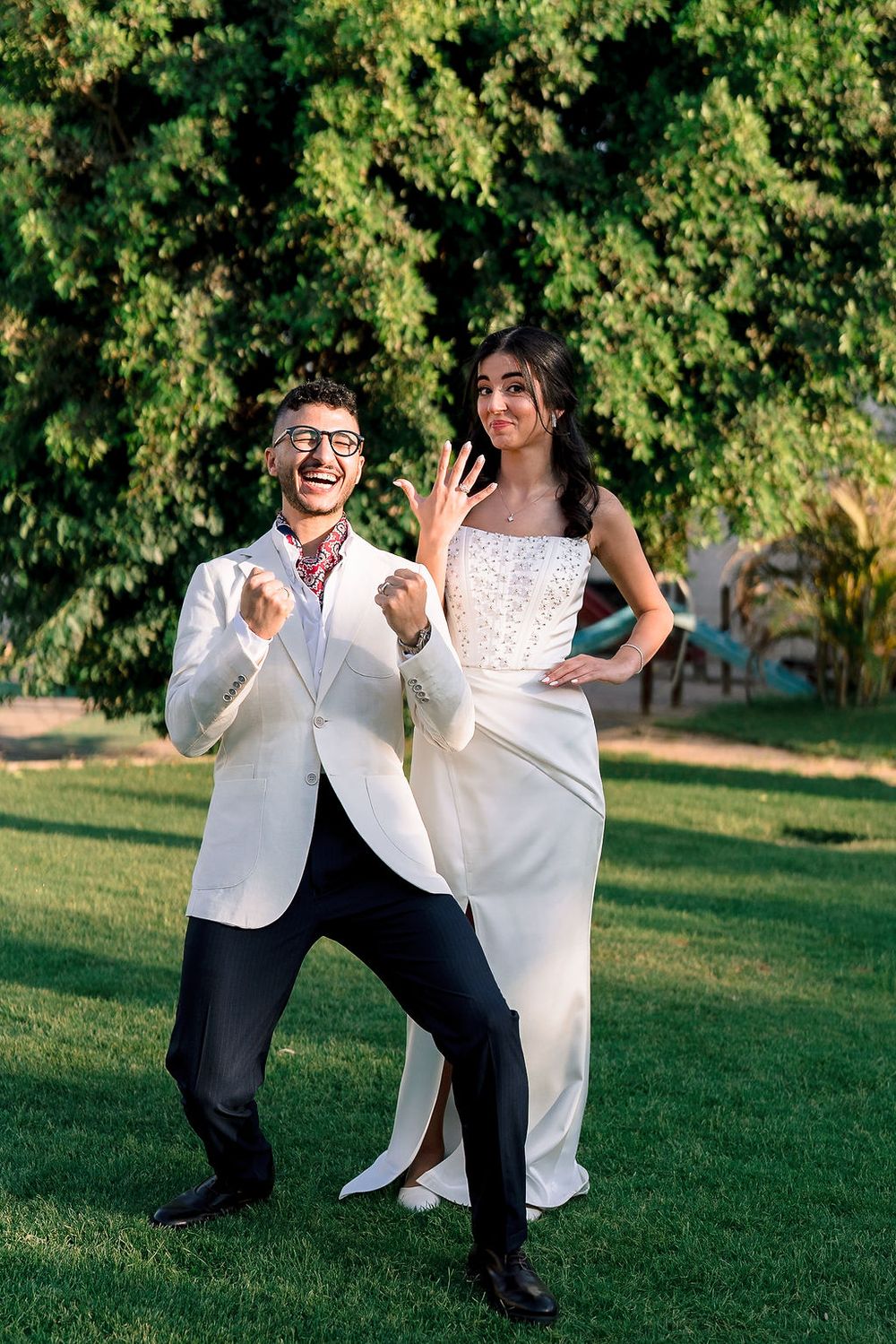 Joyful couple in wedding attire dance and pose playfully on green lawn in summer garden setting.