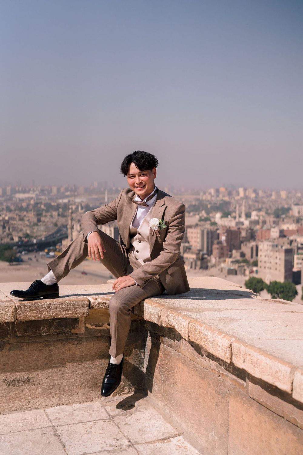 Four sequential photos show someone in formal attire reclining on a stone wall overlooking a cityscape on a sunny day.