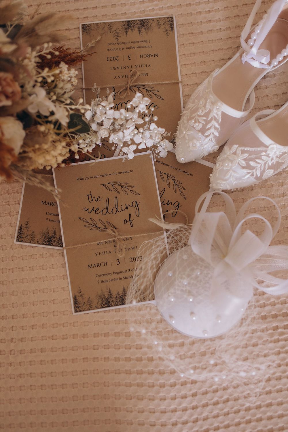 Elegant white lace wedding shoes next to rustic kraft paper wedding invitations with dried flowers on textured fabric.