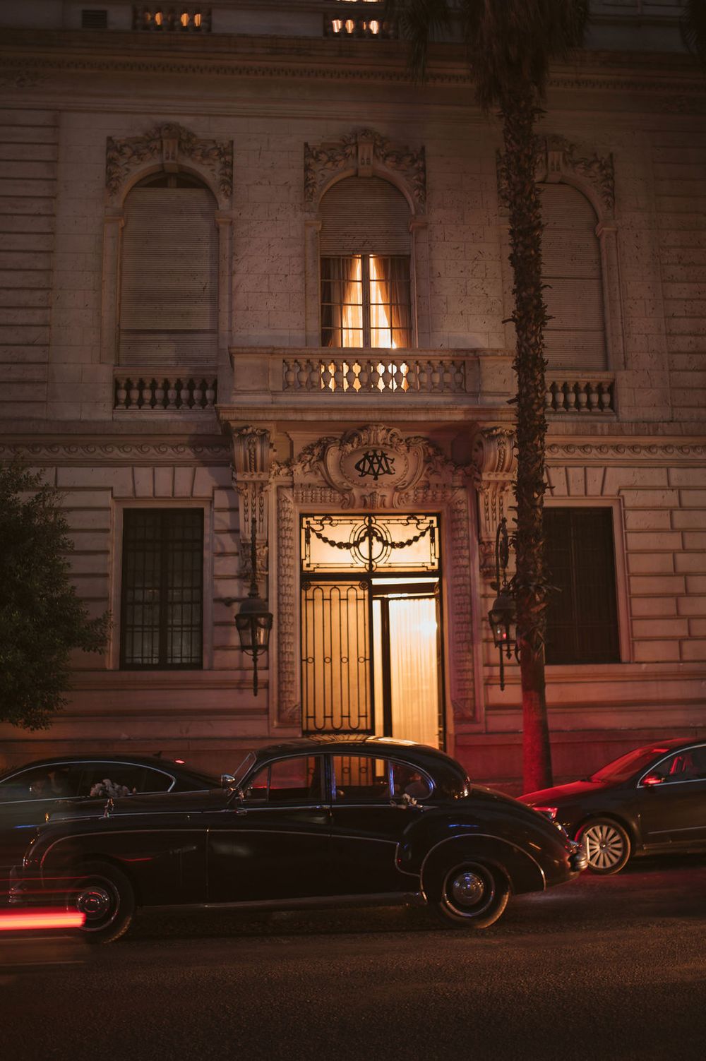 Elegant historic building facade with ornate entrance and lit balcony at night, with cars parked along dimly lit street.