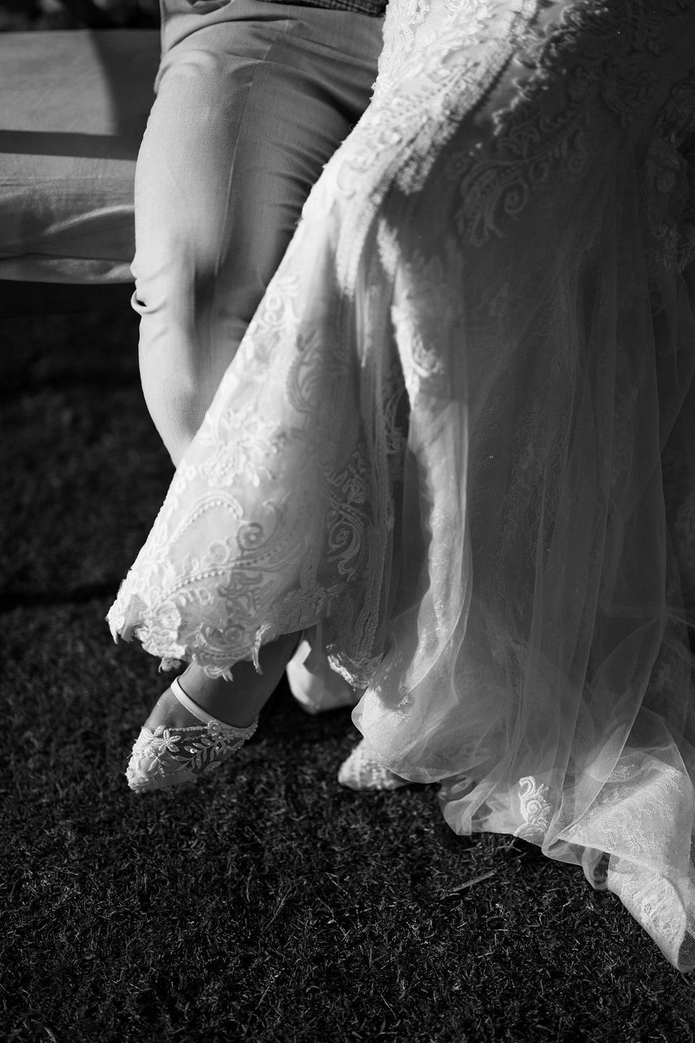 A black and white detail shot of a white lace wedding dress hem and casual shoes on grass.