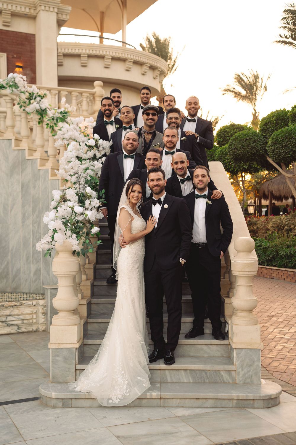 Wedding party poses in formal attire outdoors between ornate stone pillars at elegant venue.