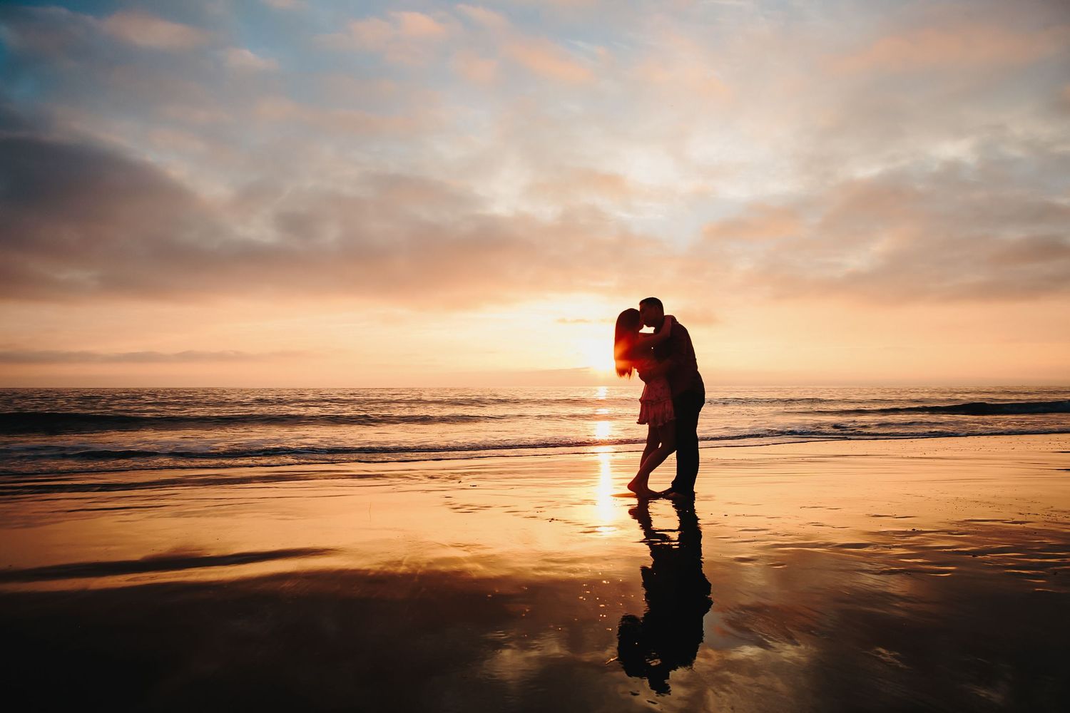 Silhouettes embrace on beach at sunset with dramatic orange sky and ocean waves creating romantic ambiance.