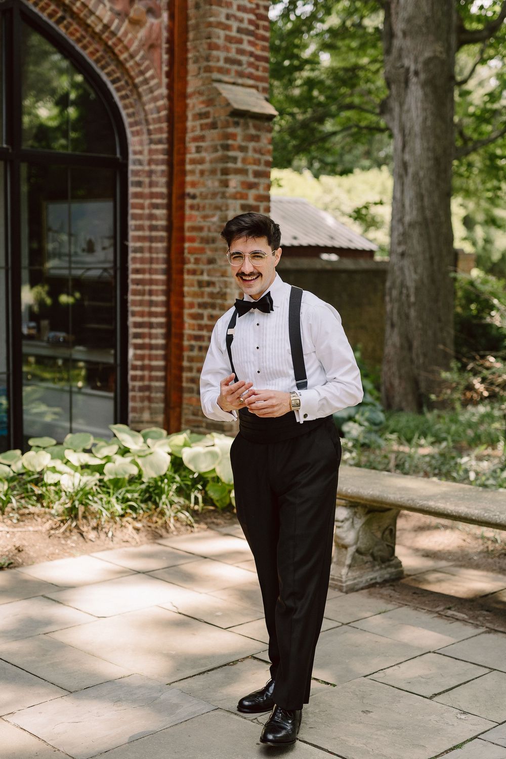 Groom in classic black tuxedo and bowtie poses in front of brick building with arched windows.