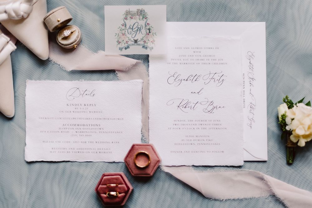 Elegant white wedding invitation suite with rose gold rings in hexagonal box and soft pink silk ribbons displayed on blue fabric.
