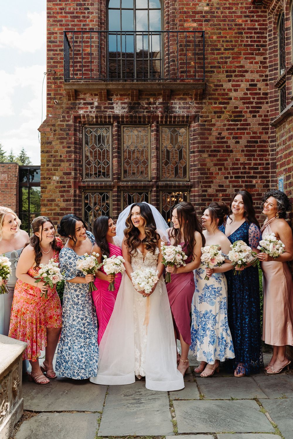 Bridal party in colorful floral dresses poses together against rustic brick building.