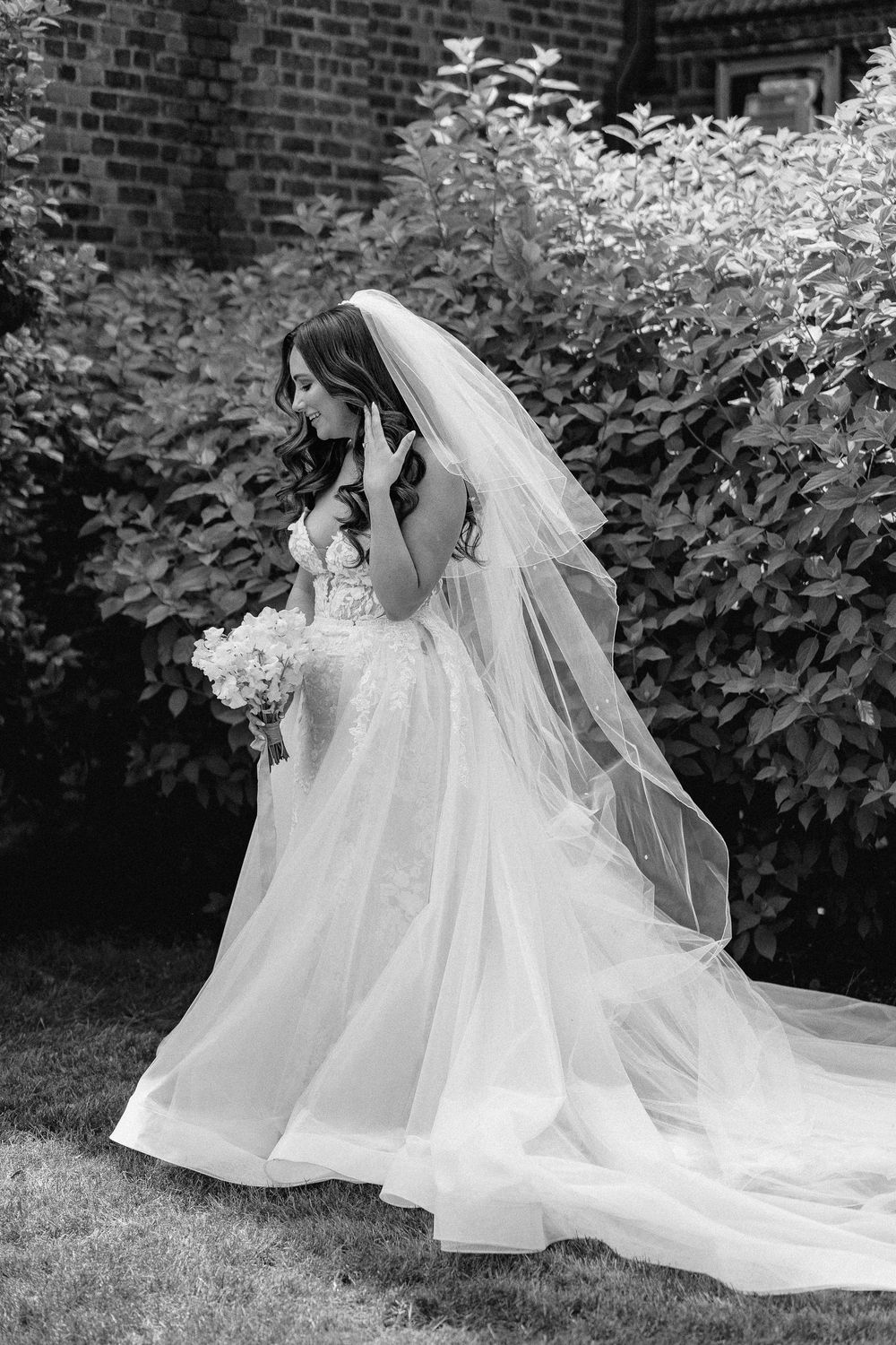 A bride in a flowing white ballgown and long veil stands against a backdrop of lush green foliage and brick wall.