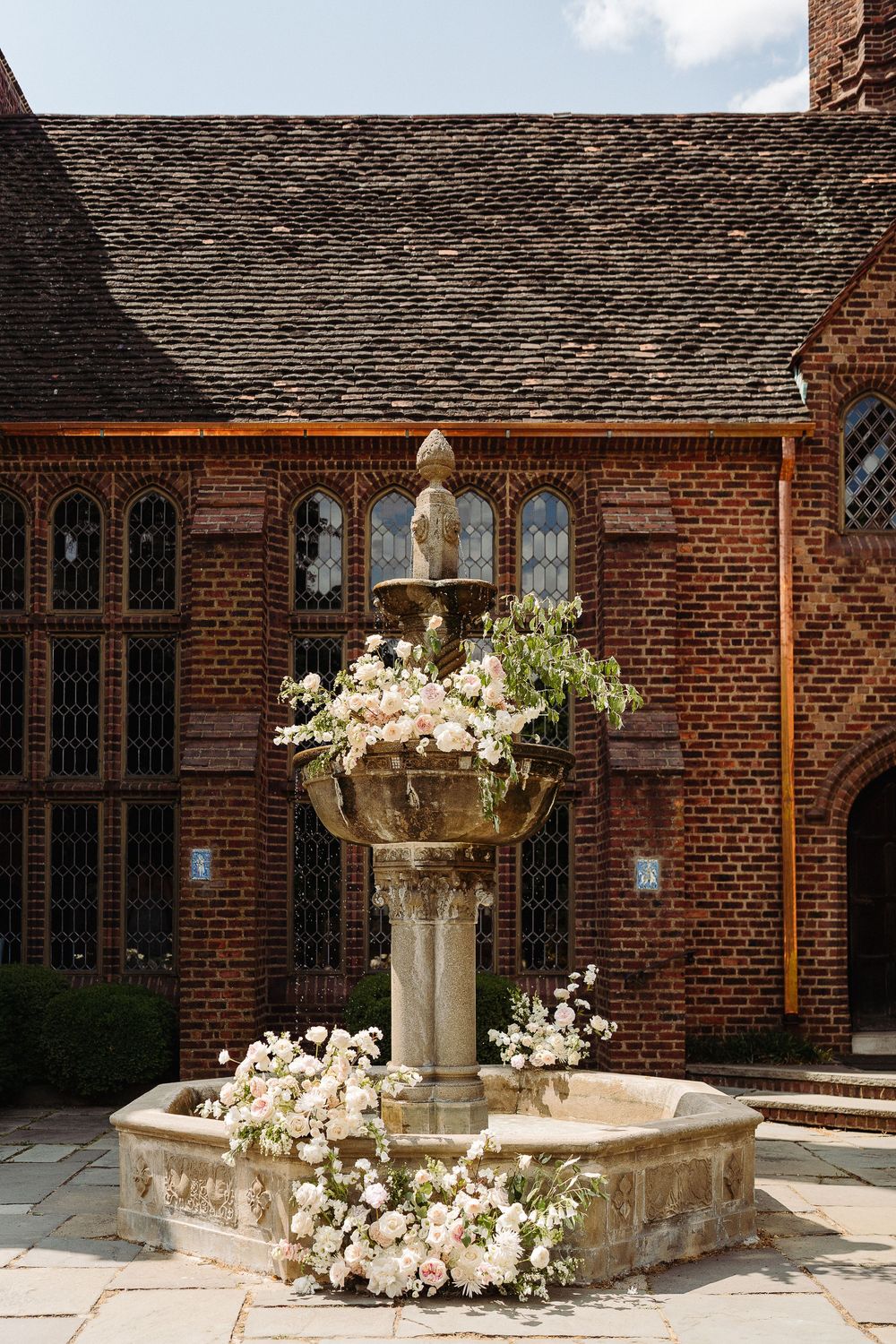 Stone fountain decorated with white flowers and greenery outside a brick church building with arched windows.