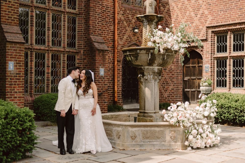 Wedding couple walks along historic brick building with white cherry blossoms framing the romantic scene.