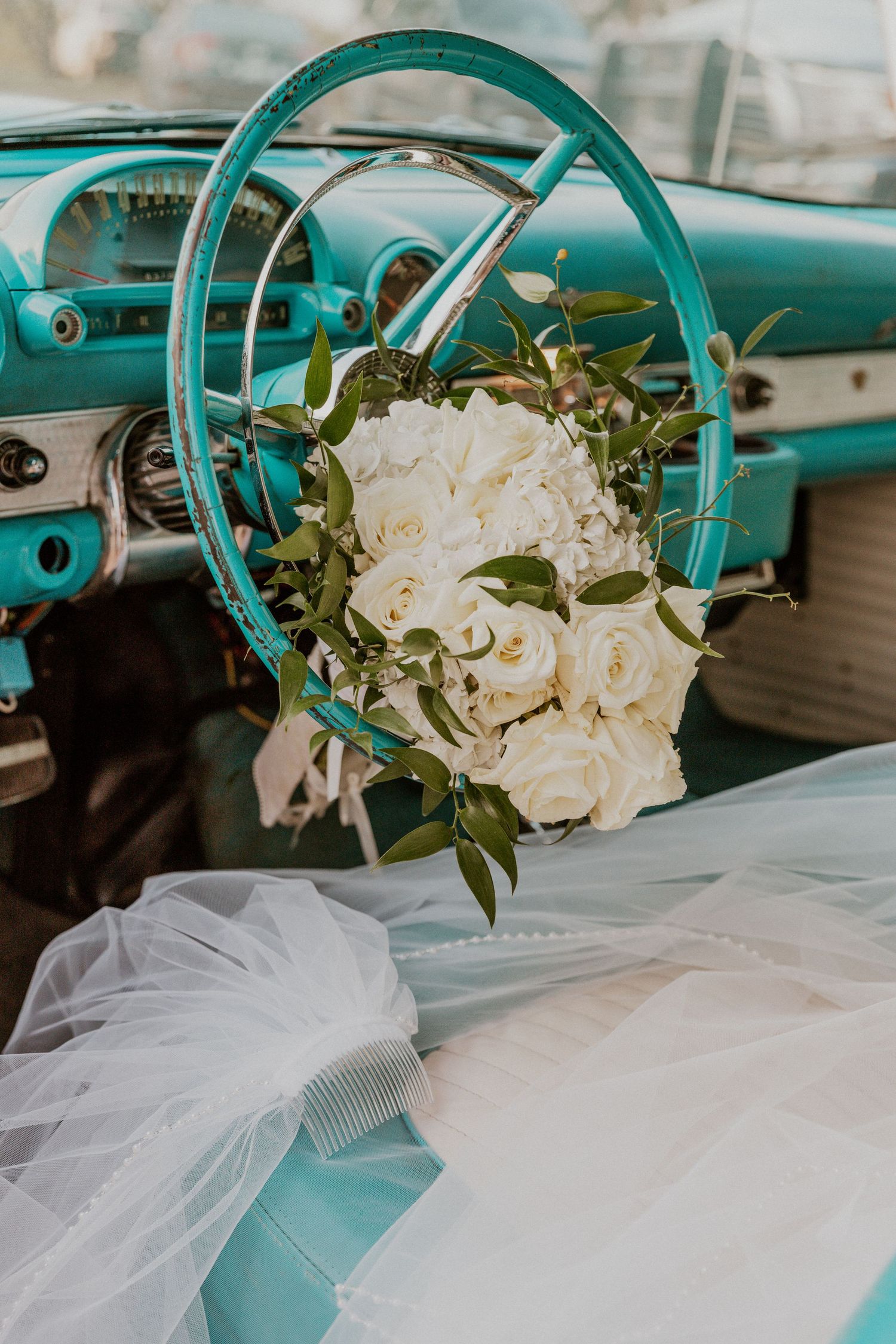 White rose bouquet sitting in a vintage car's steering wheel with a wedding veil laid across the seat.