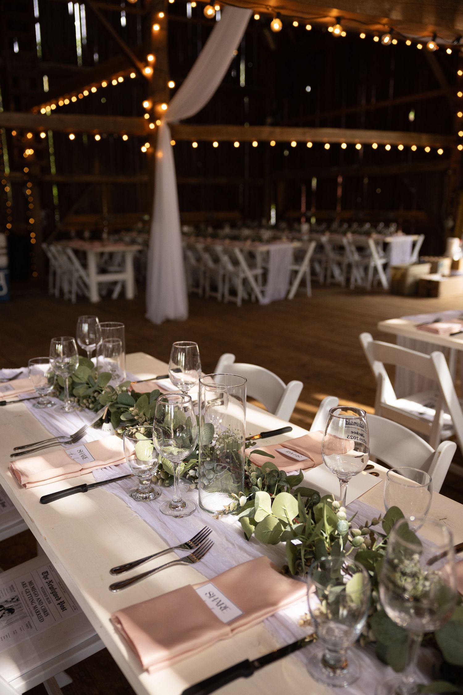 Elegant wedding reception table settings with white linens, greenery centerpieces and string lights overhead in rustic venue.