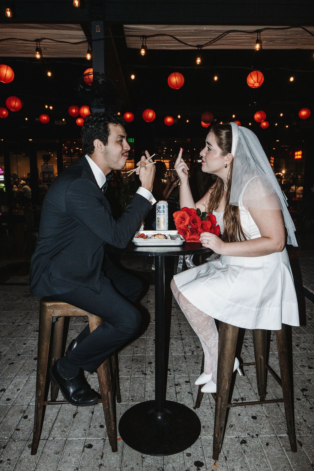 A newlywed couple shares milkshakes while sitting at a high table decorated with red paper lanterns.