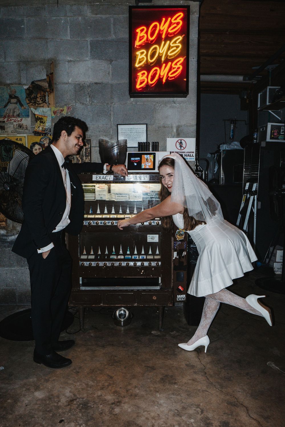 A couple in wedding attire dances together in front of a neon sign in a dimly lit industrial space.