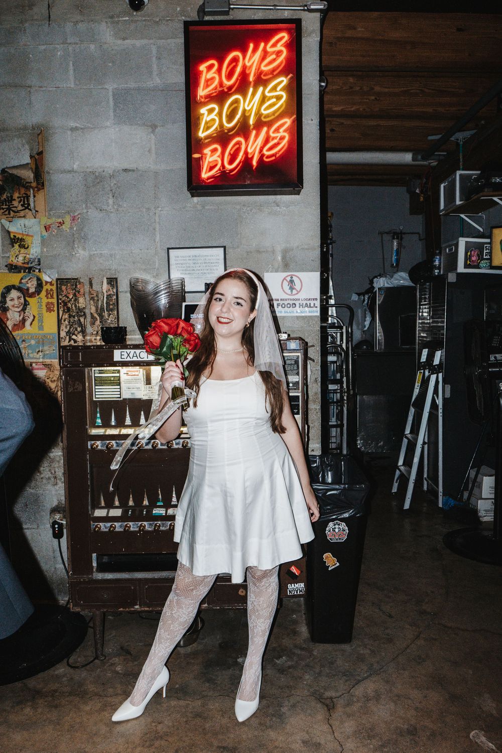 Four photos show someone in a white dress staging playful poses under a red neon 'BOYS' sign in a dark industrial space.
