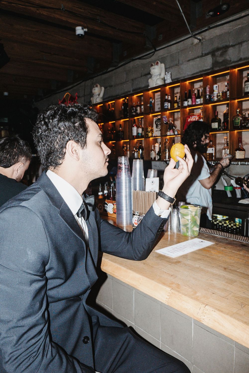 A person in a suit sits at a bar counter lined with illuminated bottles, raising a drink in their hand.