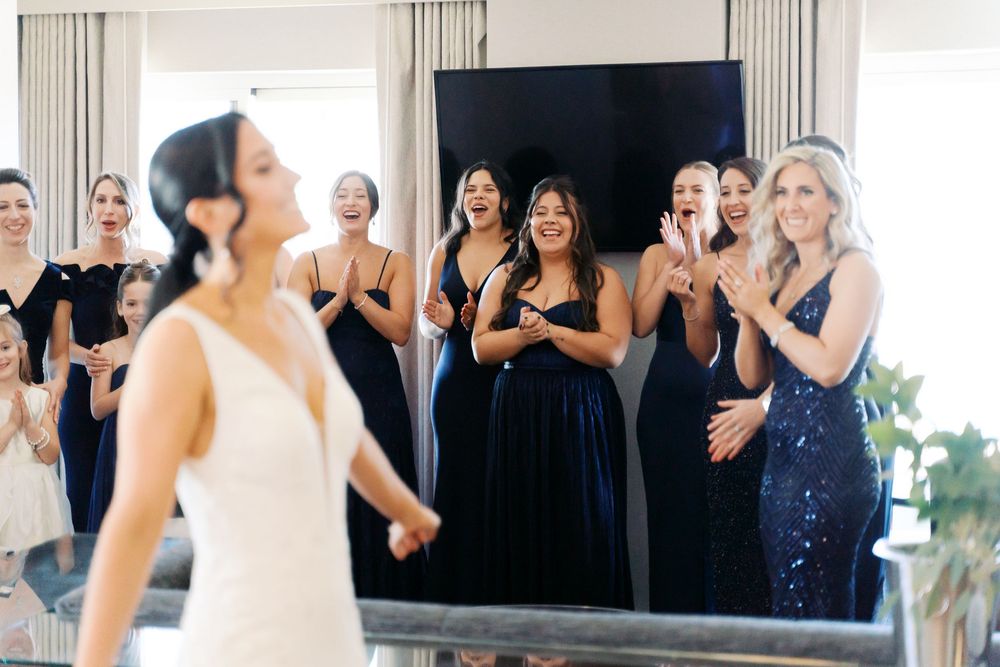 Guests in formal attire stand in rows for a wedding reception photo in a bright room with white curtains.