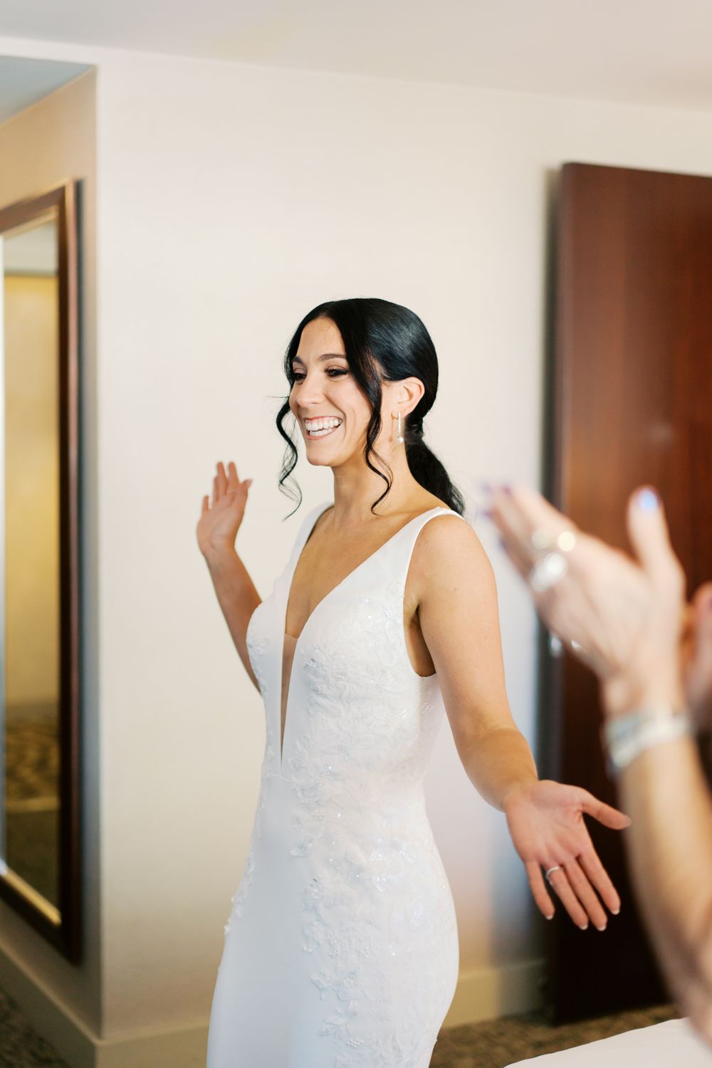 A happy bride in a white gown smiles and gestures joyfully while getting ready in a bright room.