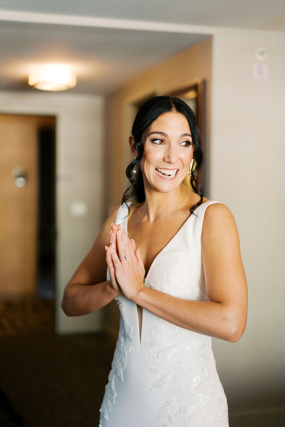 Woman in a white lace dress smiles and poses elegantly in a room with natural lighting.