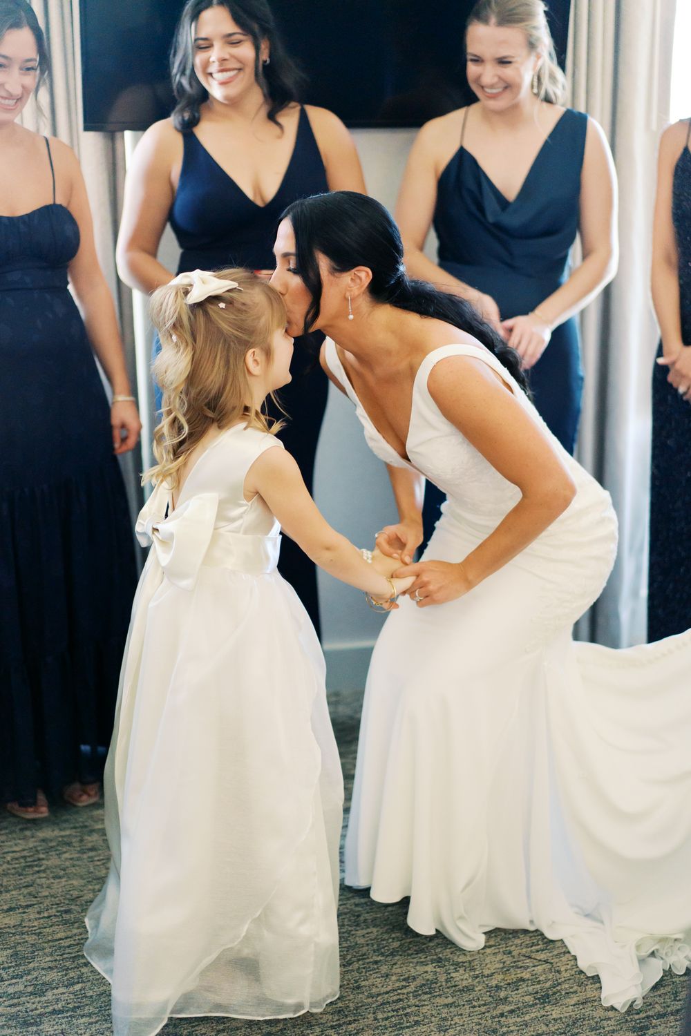 Young flower girl in white dress dances joyfully with wedding attendants in navy dresses.