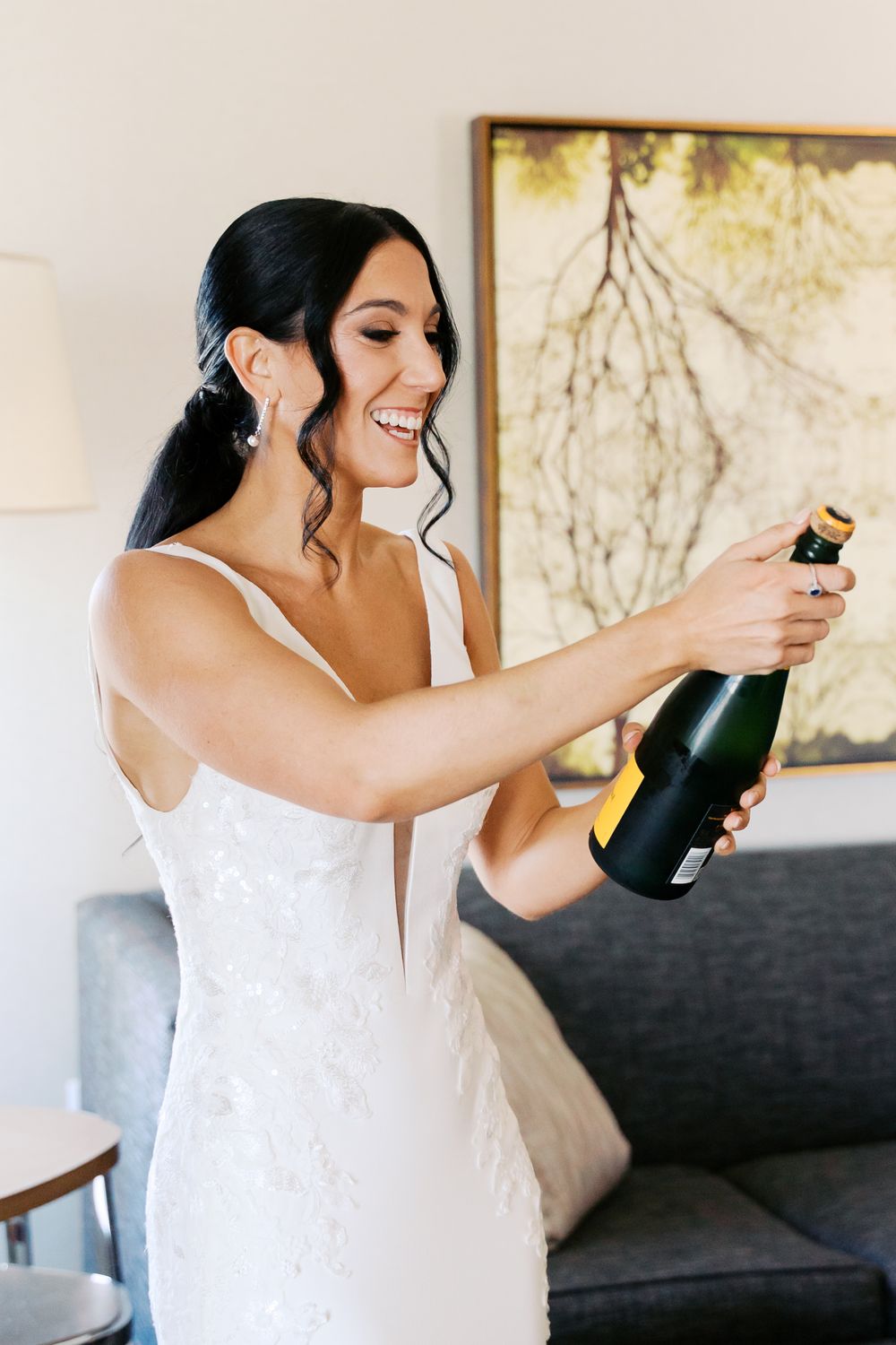 Someone in a white dress opening a champagne bottle during wedding preparations.