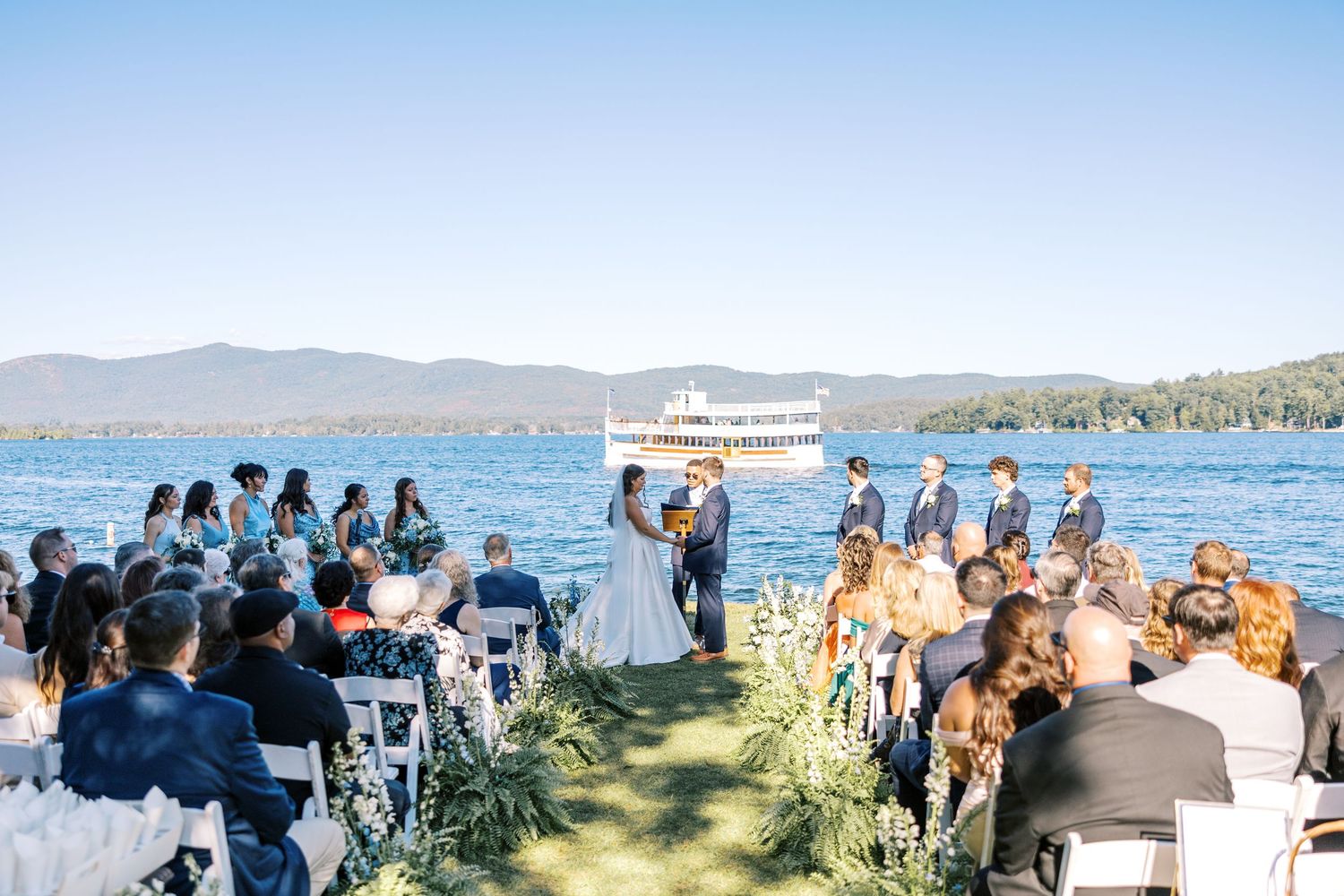 A scenic lakeside wedding ceremony with guests seated in white chairs overlooking blue waters and mountains.