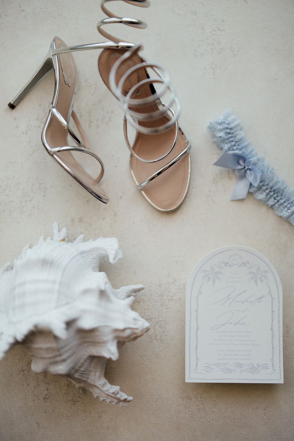 Wedding accessories including silver heels, decorative paper goods, and white feather accents on a neutral surface.