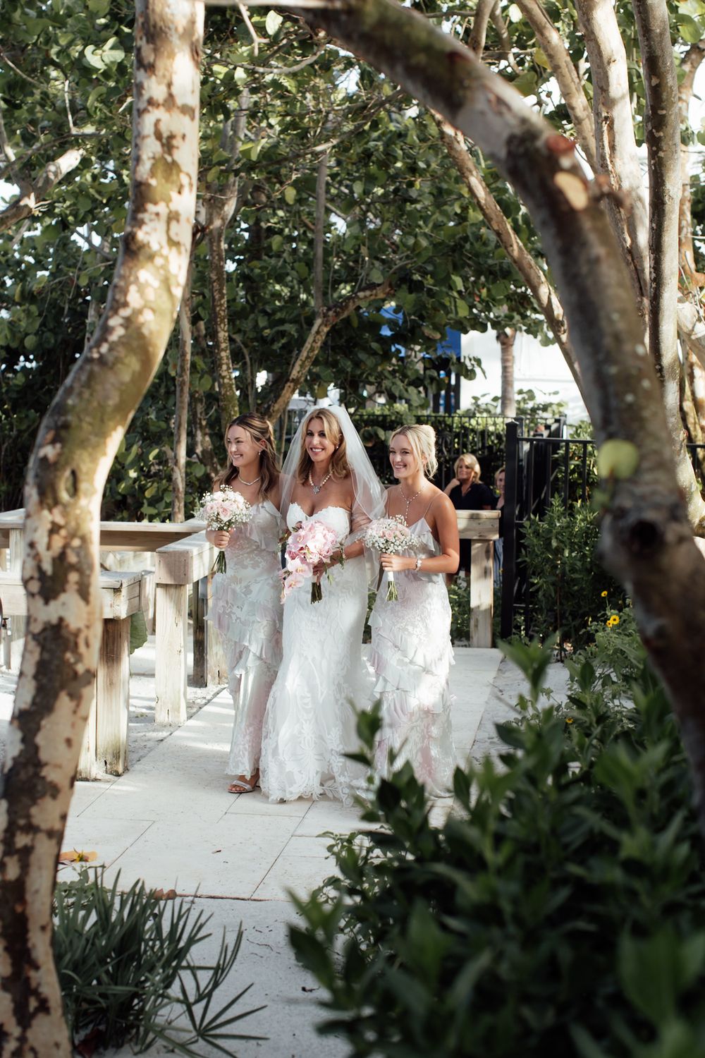 A group in white dresses poses together on a garden walkway with trees overhead.