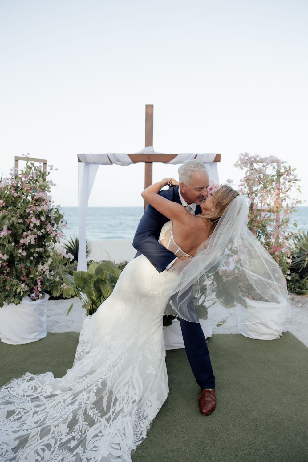 Romantic wedding ceremony dip kiss in front of a cross with ocean views and floral arrangements.