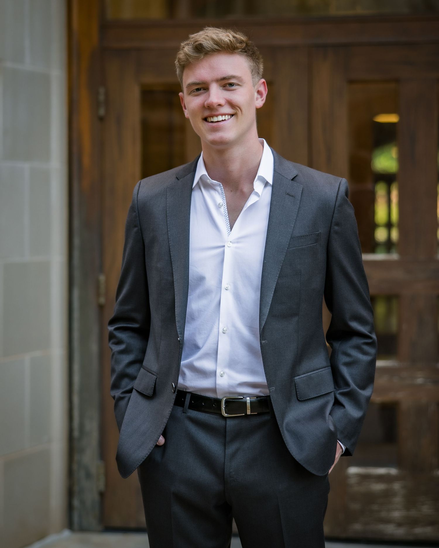 Young professional in grey suit and white shirt stands confidently against stone building exterior.