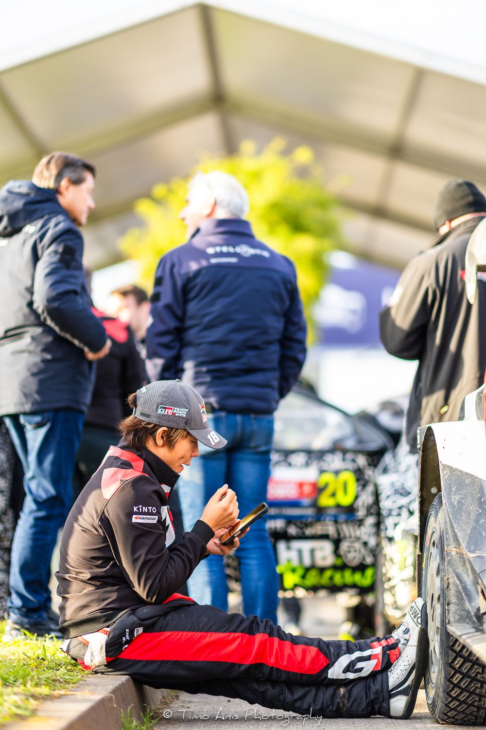 Motorsport participant sits on ground checking phone while others stand at racing event.