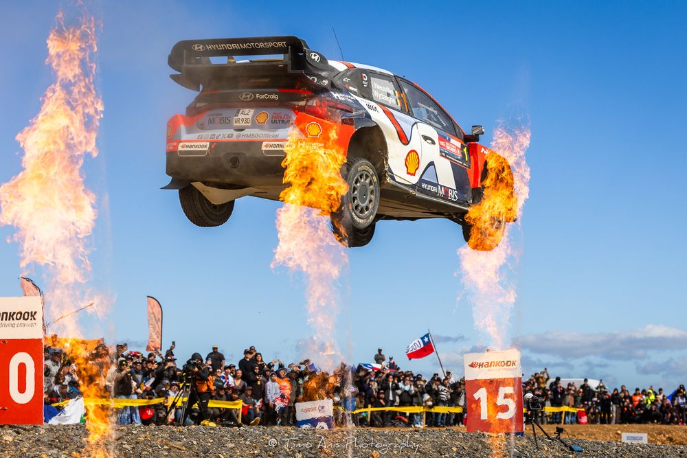 A dramatic sequence shows a car launching off a ramp and bursting into flames against a blue sky with spectators watching.
