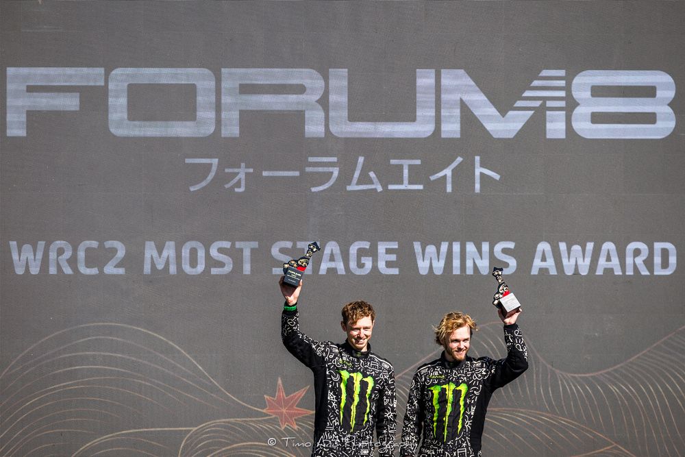 Rally car drivers celebrate on podium with energy drink sponsor banners and trophies in background.