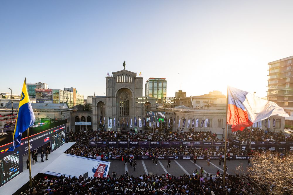 Crowd gathers at a religious site during sunset with large basilica and cathedral buildings visible in background.