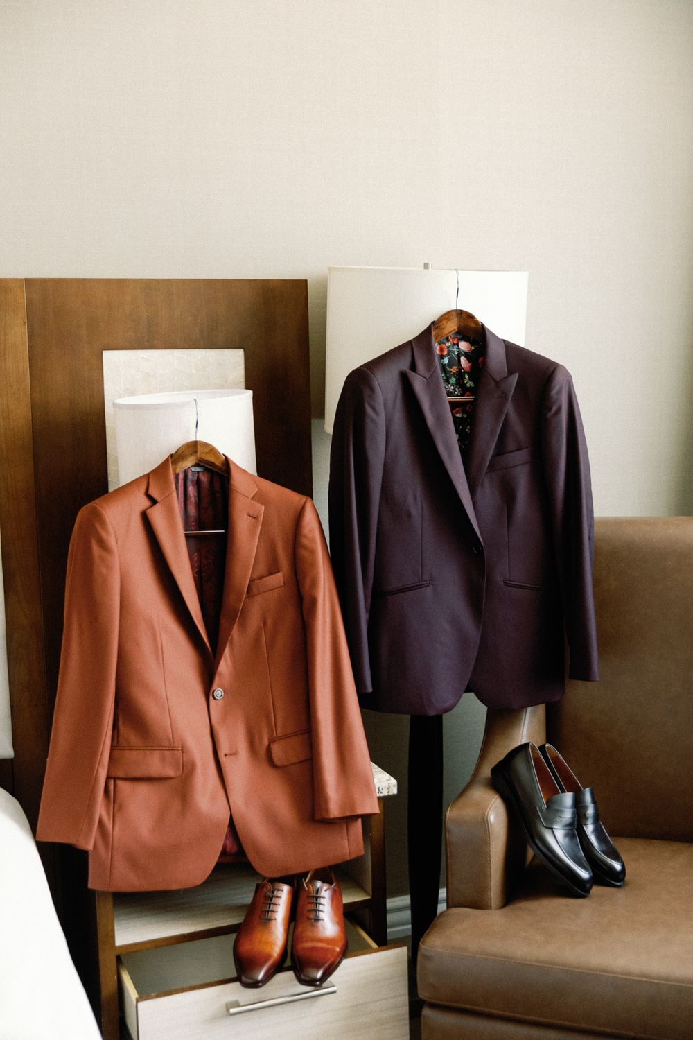 Two mens suit jackets in rust and burgundy hanging with matching dress shoes below in hotel room.