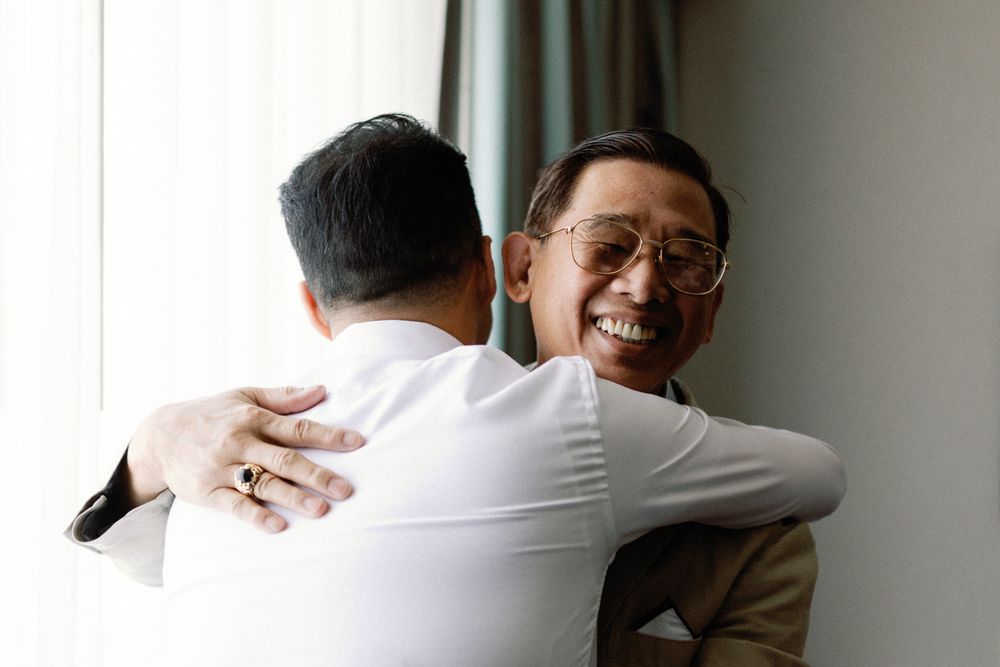 Two men share a warm embrace, one smiling joyfully, in a bright room during a wedding celebration.