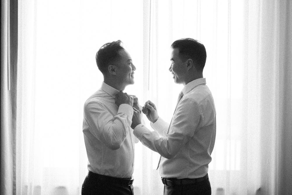 Black and white photo of two men in white shirts helping each other with tie before wedding ceremony.