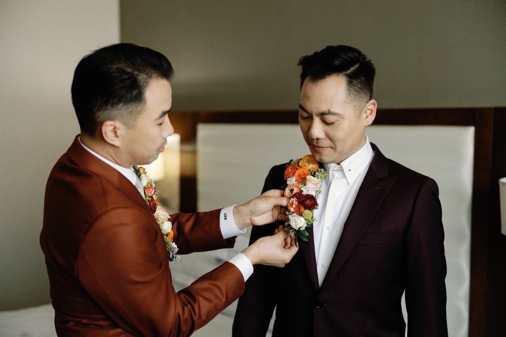 Two grooms in burgundy suits pin boutonnieres on each other while preparing for their wedding.