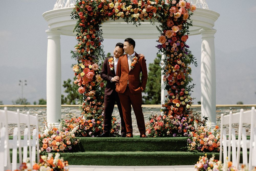Two grooms stand together under a stunning floral arch of orange and pink blooms at outdoor wedding.