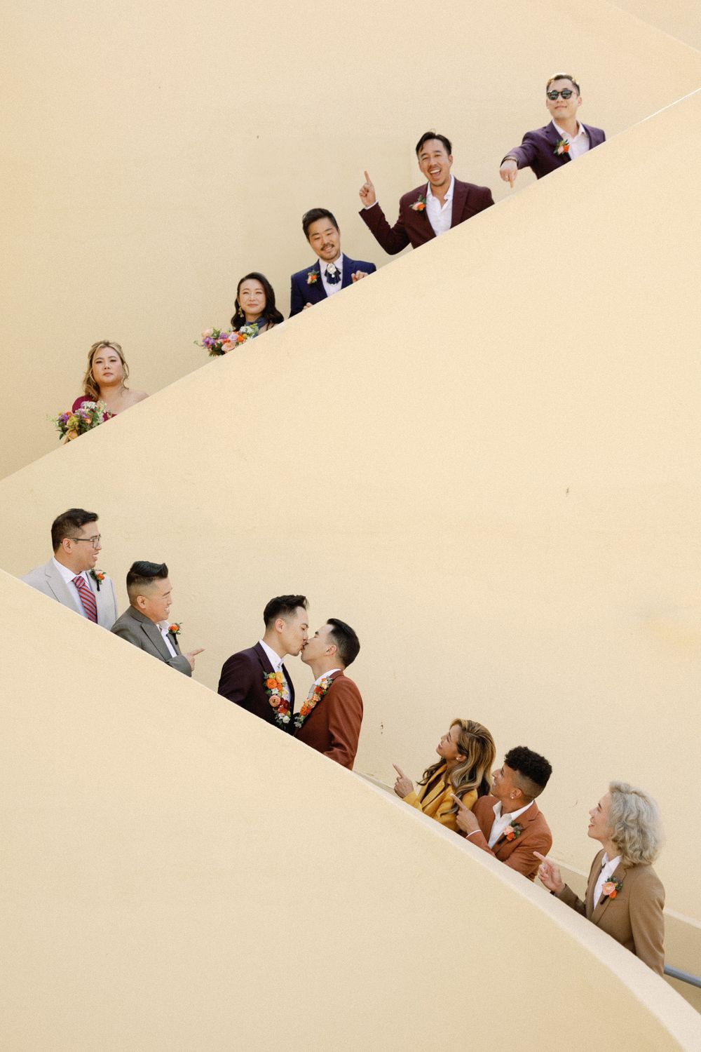 Wedding party poses on a curved staircase as two grooms share a kiss during their ceremony.