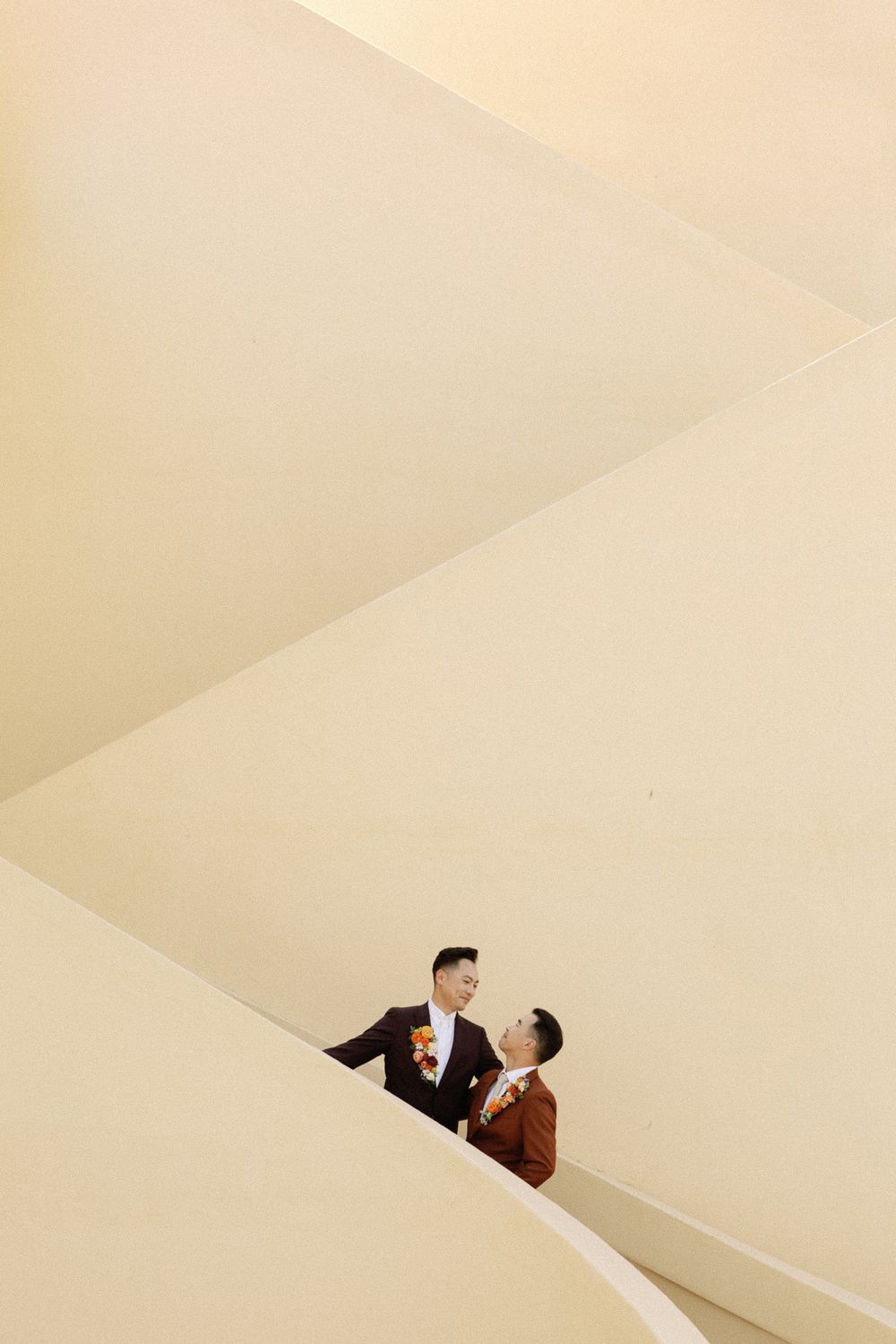 Two grooms in matching brown suits share a romantic look on geometric cream-colored stairs.