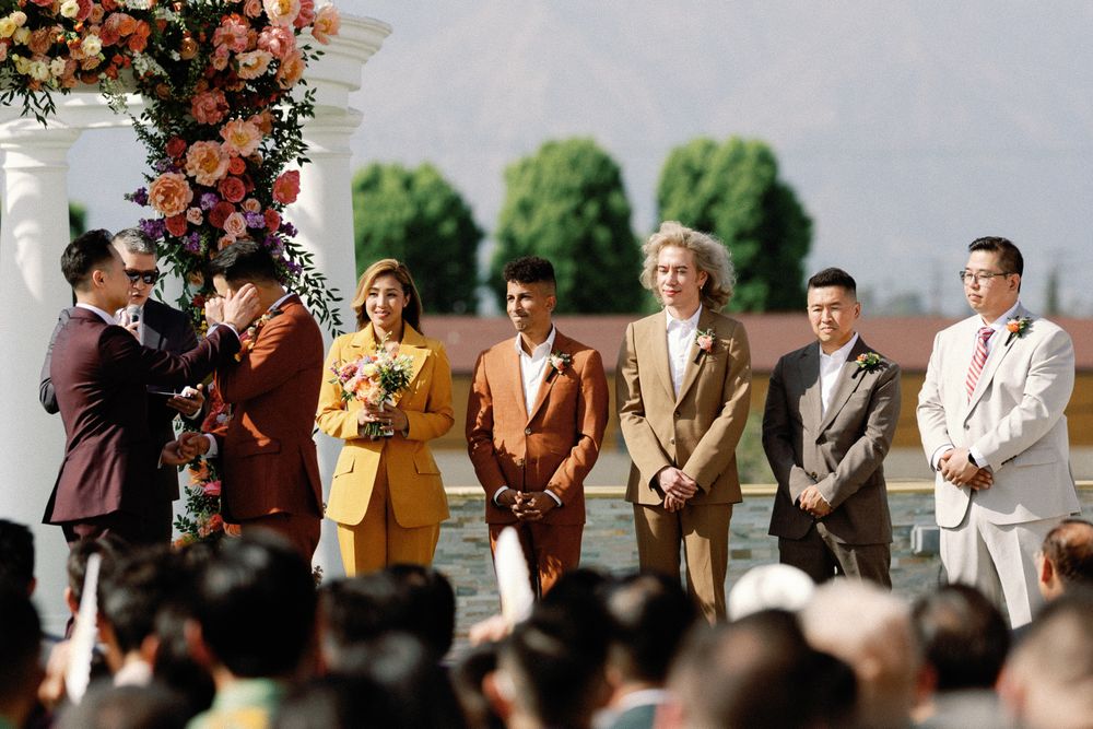 Wedding party in colorful suits stands at floral altar during outdoor same-sex wedding ceremony.