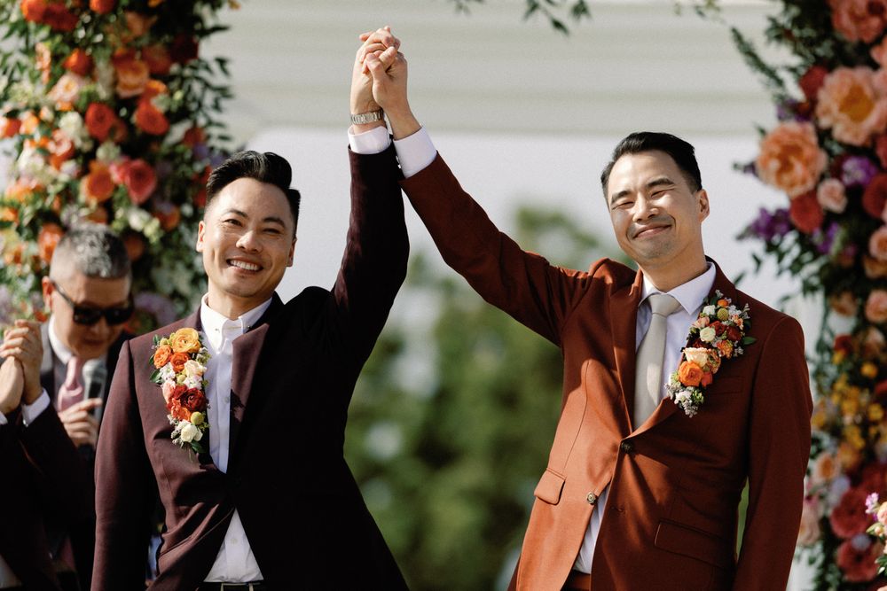 Two grooms raise joined hands triumphantly before cheering guests at their outdoor wedding ceremony.
