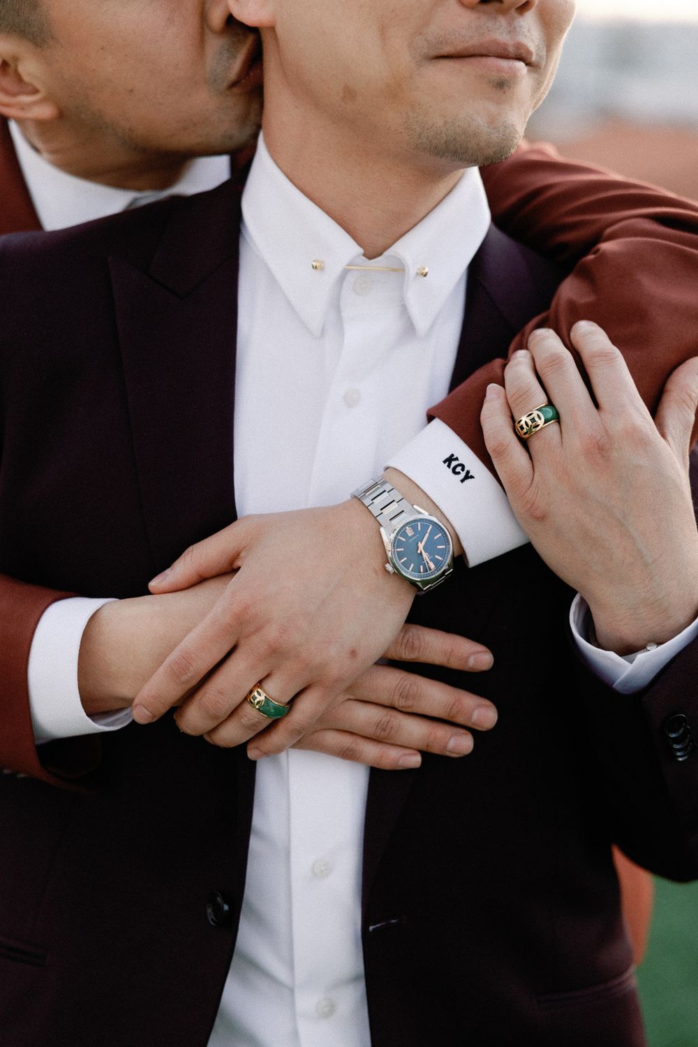 Two grooms embrace closely, one wearing a silver watch and personalized cufflinks, both in elegant dark suits.