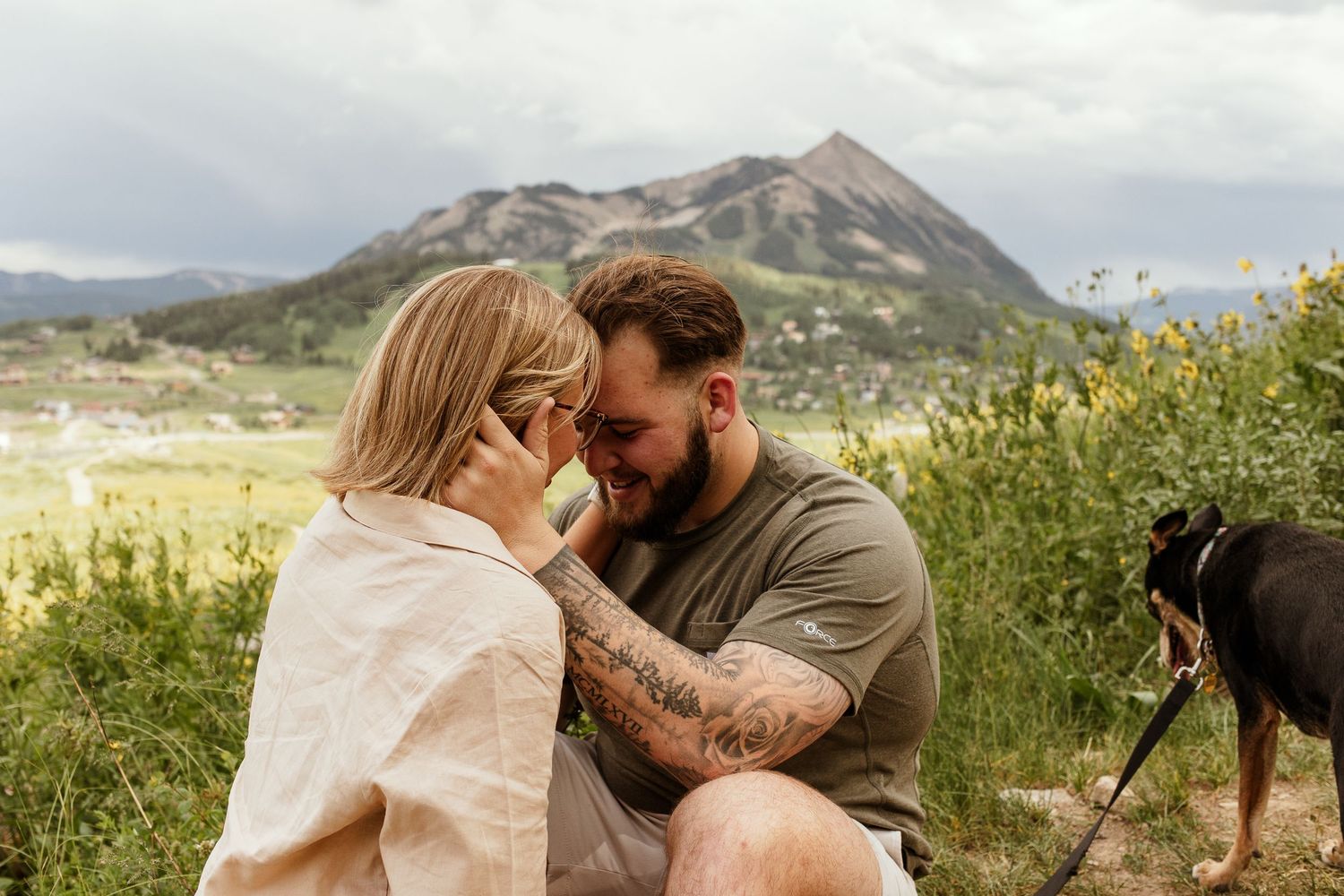 Couple embracing in a picturesque mountain landscape with wildflowers and natural scenery in the background.