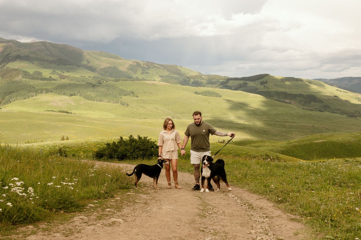 People and dogs enjoying a scenic hike along rolling green hills on a cloudy summer day in the mountains.