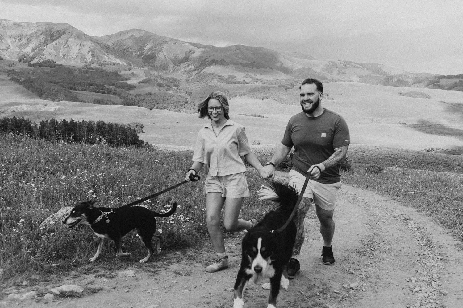 Game hunters with rifles traverse rugged mountain terrain with their hunting dogs in a black and white landscape.