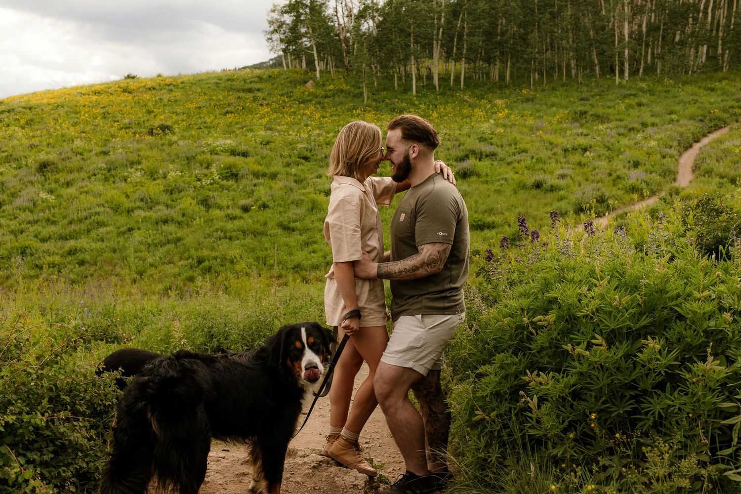 Adventurous couple sits on rocky outcrop overlooking vibrant green valley with distant trees and mountains.