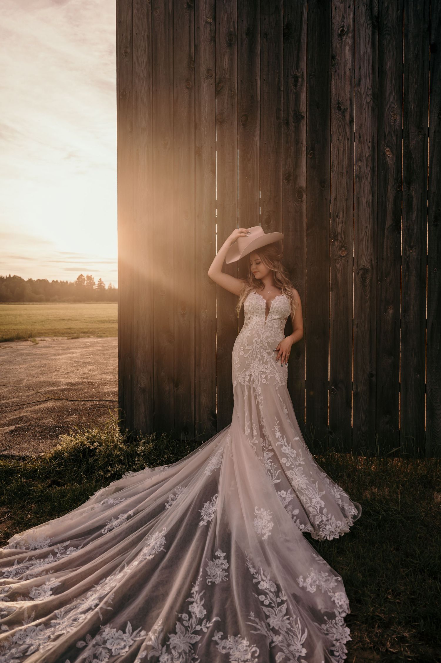 A stunning lace wedding dress with long train photographed against rustic barn wall during golden sunset.