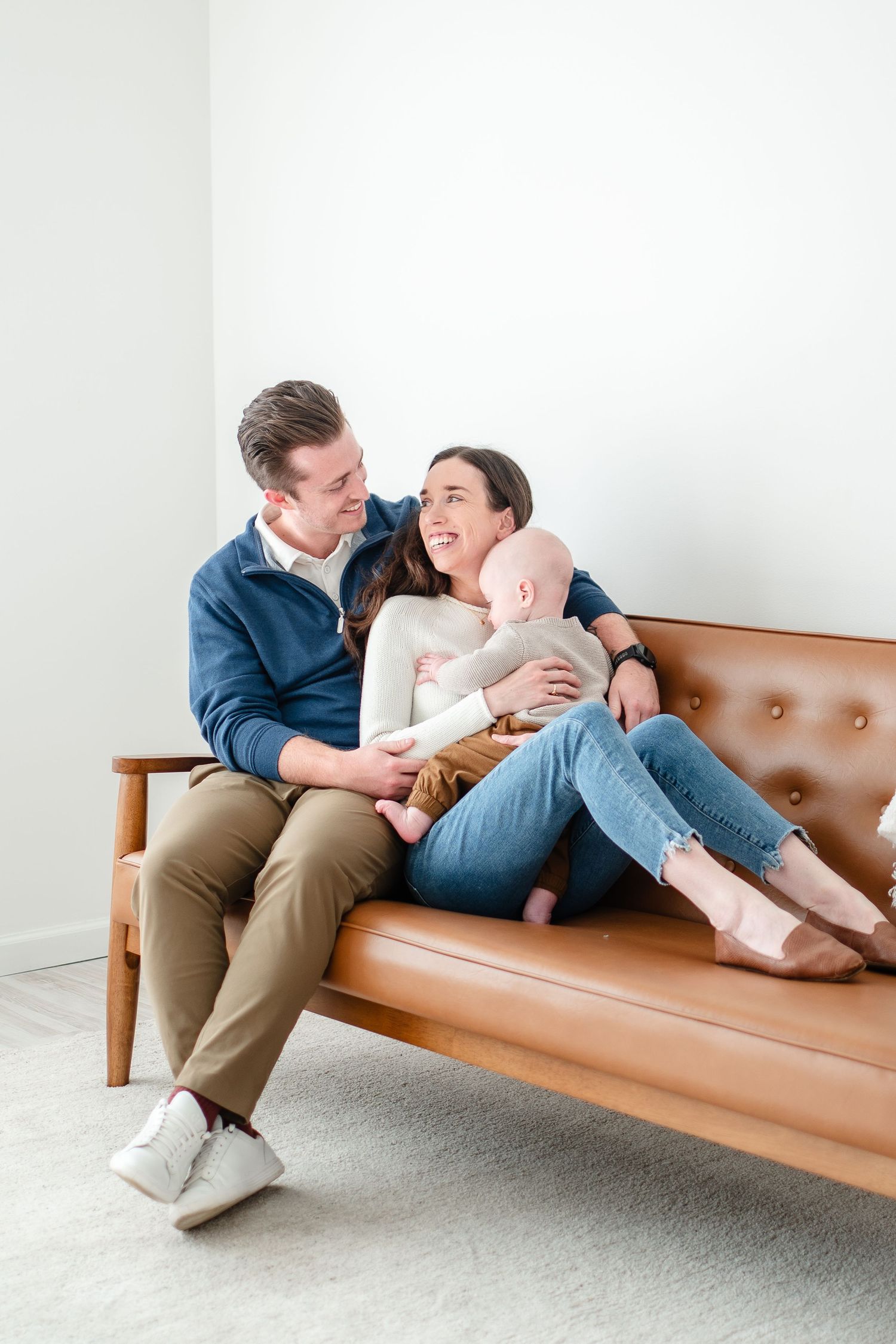 A couple reclines playfully together on a tan leather mid-century modern sofa in a bright minimalist setting.
