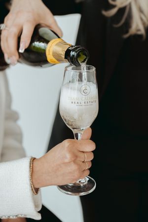 Hands pouring champagne into branded real estate company glasses.
